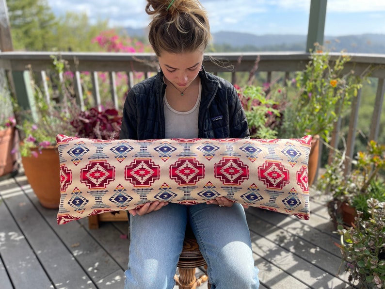 May include: A long, rectangular pillow with a geometric pattern in shades of red, blue, and white. The pillow is made of a soft, woven fabric and has a fringe trim.