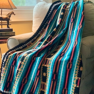 May include: A turquoise, black, red, and gold patterned fleece throw blanket with a black and white crocheted border. The blanket is draped over a beige armchair.