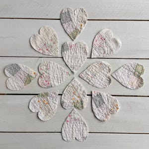 Puede incluir: Disposición de doce corazones de tela acolchada en varios patrones y colores, incluyendo blanco, rosa y gris. Los corazones están dispuestos sobre una superficie de madera blanca, creando una exhibición decorativa. Cada corazón mide aproximadamente 10 cm de ancho.