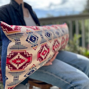 May include: A long, rectangular pillow with a cream background and a geometric pattern in shades of red, blue, and yellow. The pillow is made of fabric and has a fringed edge.