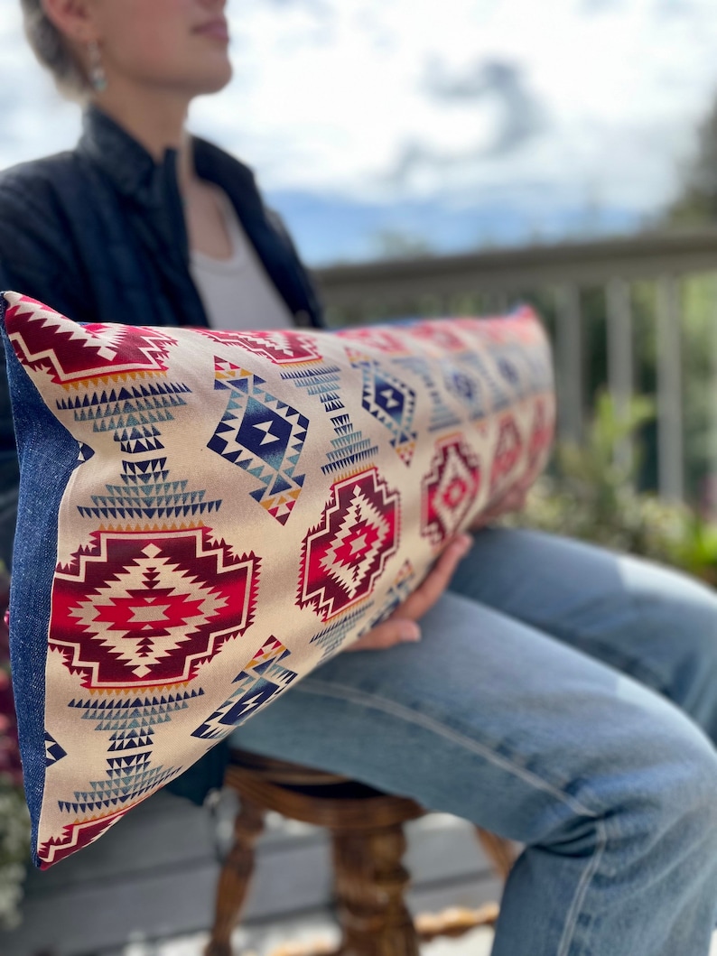 May include: A long, rectangular pillow with a cream background and a geometric pattern in shades of red, blue, and yellow. The pillow is made of fabric and has a fringed edge.