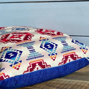 May include: A square throw pillow with a geometric pattern in shades of red, blue, and white on a cream background. The pillow has a blue denim border.