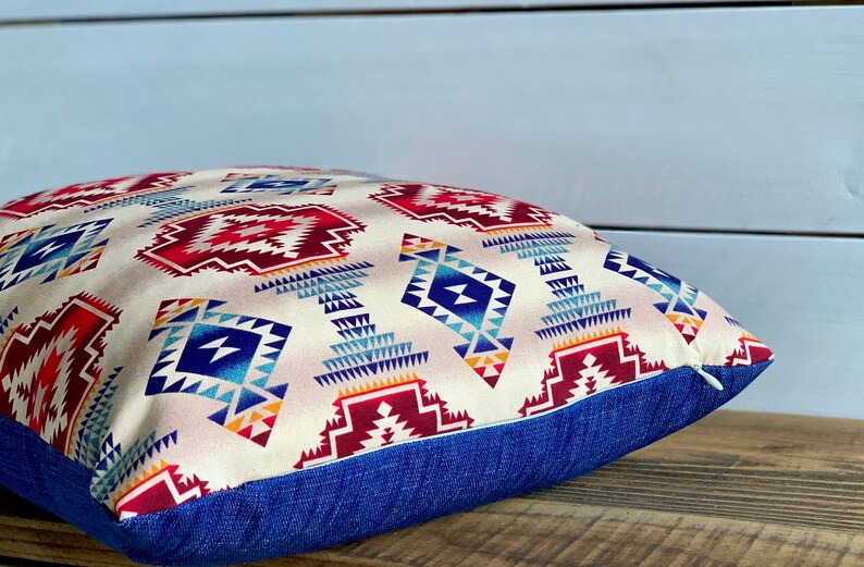 May include: A square throw pillow with a geometric pattern in shades of red, blue, and white on a cream background. The pillow has a blue denim border.