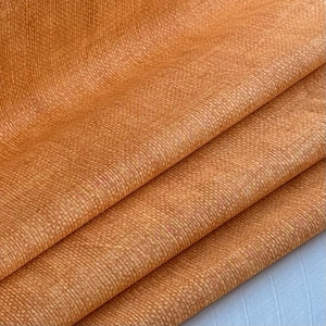 Puede incluir: Primer plano de una tela naranja texturizada con un brillo sutil. La tela está doblada en tres partes, revelando la textura y el color.