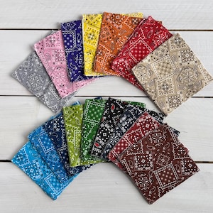 May include: A collection of colorful bandanas in various shades, including blue, pink, yellow, orange, red, and brown. Each bandana features a classic paisley pattern. The bandanas are arranged on a white wooden surface.