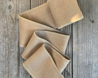 May include: A roll of natural brown burlap ribbon with a white stitched edge. The ribbon is rolled up and lying on a wooden surface.
