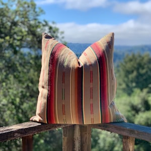 May include: A decorative pillow with a striped pattern in shades of brown, red, and green. The pillow is rectangular and appears to be made of woven fabric. It is resting on a wooden railing outdoors.