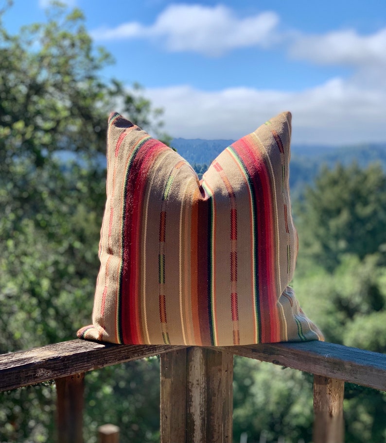 May include: A decorative pillow with a striped pattern in shades of brown, red, and green. The pillow is rectangular and appears to be made of woven fabric. It is resting on a wooden railing outdoors.