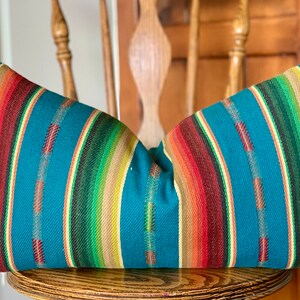May include: A teal blue pillow with colorful stripes in shades of red, orange, yellow, green, and brown. The pillow is made of a woven fabric and has a soft, comfortable texture.