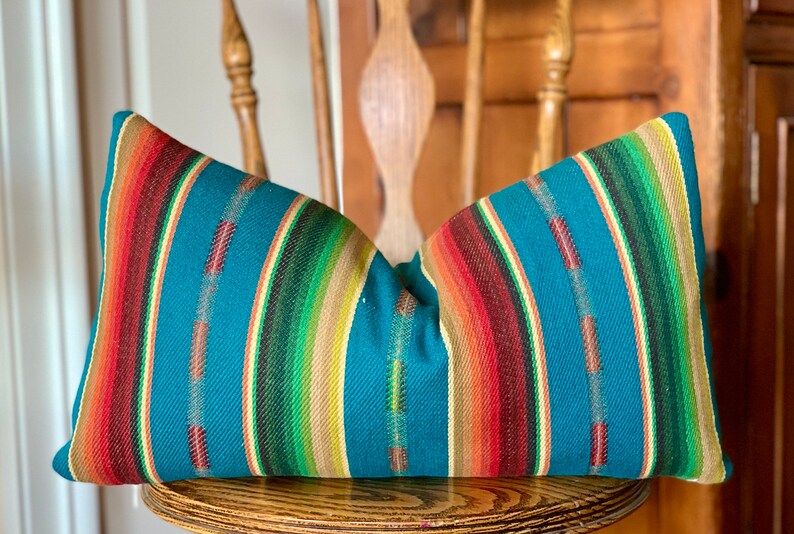May include: A teal blue pillow with colorful stripes in shades of red, orange, yellow, green, and brown. The pillow is made of a woven fabric and has a soft, comfortable texture.