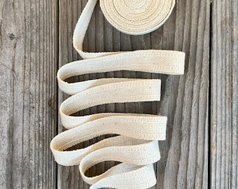 May include: A roll of off-white cotton webbing, a type of fabric tape, is coiled on a wooden surface. The webbing is woven with a subtle texture.