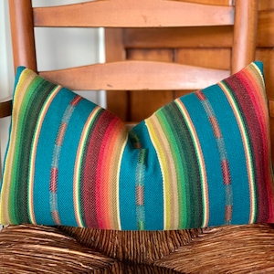 May include: A teal blue pillow with colorful stripes in shades of red, orange, yellow, green, and brown. The pillow is sitting on a wooden chair.