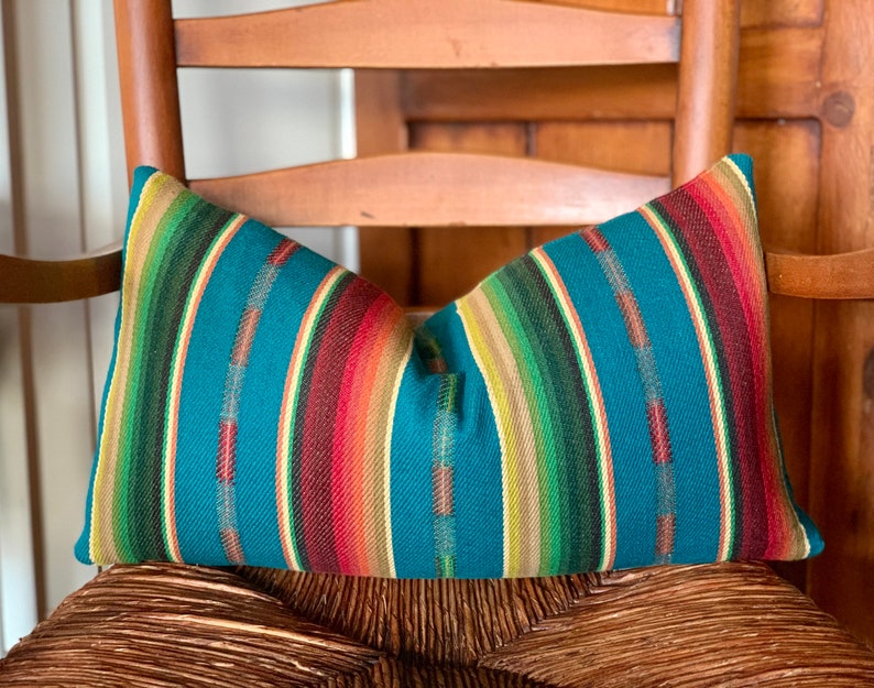 May include: A teal blue pillow with colorful stripes in shades of red, orange, yellow, green, and brown. The pillow is sitting on a wooden chair.
