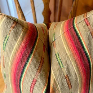 May include: Close-up of a decorative pillow with a striped pattern. The pillow features a tan background with vertical stripes in red, green, brown, and white. The pillow is placed on a wooden chair.