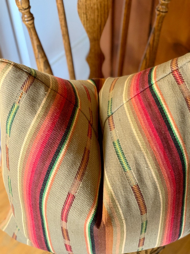 May include: Close-up of a decorative pillow with a striped pattern. The pillow features a tan background with vertical stripes in red, green, brown, and white. The pillow is placed on a wooden chair.