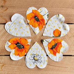 May include: Heart-shaped fabric pieces with floral patterns. Some hearts feature orange poppies with black centers, while others have small yellow daisies on a white background. The pieces are arranged on a wooden surface.