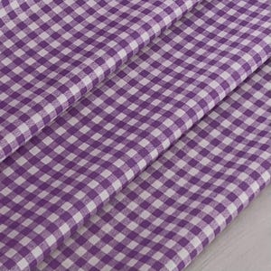 May include: A close-up of a purple and white gingham fabric. The fabric is folded, showing the pattern at different angles. The pattern is a classic checkered design, with squares of purple and white.