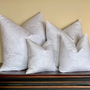 May include: Five white down pillows of varying sizes arranged on a dark wooden surface. The pillows are all made of white down and have a soft, fluffy texture.