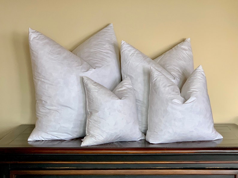 May include: Five white down pillows of varying sizes arranged on a dark wooden surface. The pillows are all made of white down and have a soft, fluffy texture.