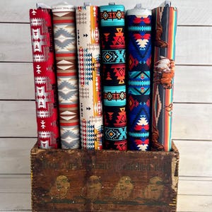 May include: Six rolls of fabric with colorful geometric patterns in shades of red, blue, yellow, and gray. The fabric is displayed in a wooden crate with a painted design.