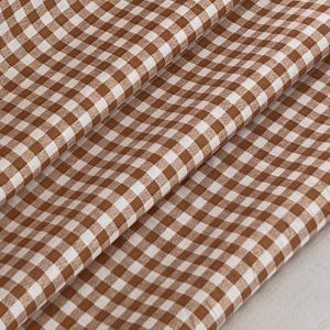 Może przedstawiać: Brązowo-biała tkanina w kratę z wzorem gingham. Tkanina jest złożona, tworząc cienie i głębię. Wzór to klasyczny design, odpowiedni do różnych projektów rzemieślniczych i dekoracji wnętrz.
