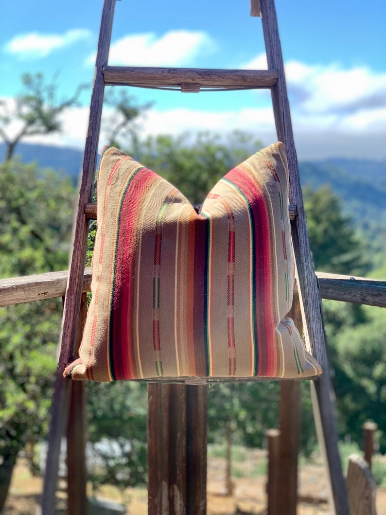 May include: A decorative pillow with a striped pattern in shades of brown, red, and green. The pillow is displayed on a wooden ladder outdoors, with a backdrop of trees and a blue sky.