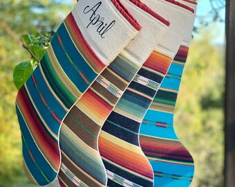 Könnte beinhalten: Vier bunte gestreifte Strümpfe mit rotem Besatz, die an einem Holzgeländer hängen. Der oberste Strumpf ist mit dem Namen "April" personalisiert.
