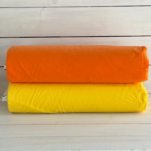 May include: Two rolls of fabric, one bright orange and one yellow, are stacked on a white wooden surface. The fabric appears to be a soft material, possibly cotton or a similar textile. The rolls are neatly folded.