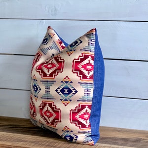 May include: A decorative pillow with a geometric pattern in shades of red, blue, and yellow on a cream background. The pillow has a denim blue border.