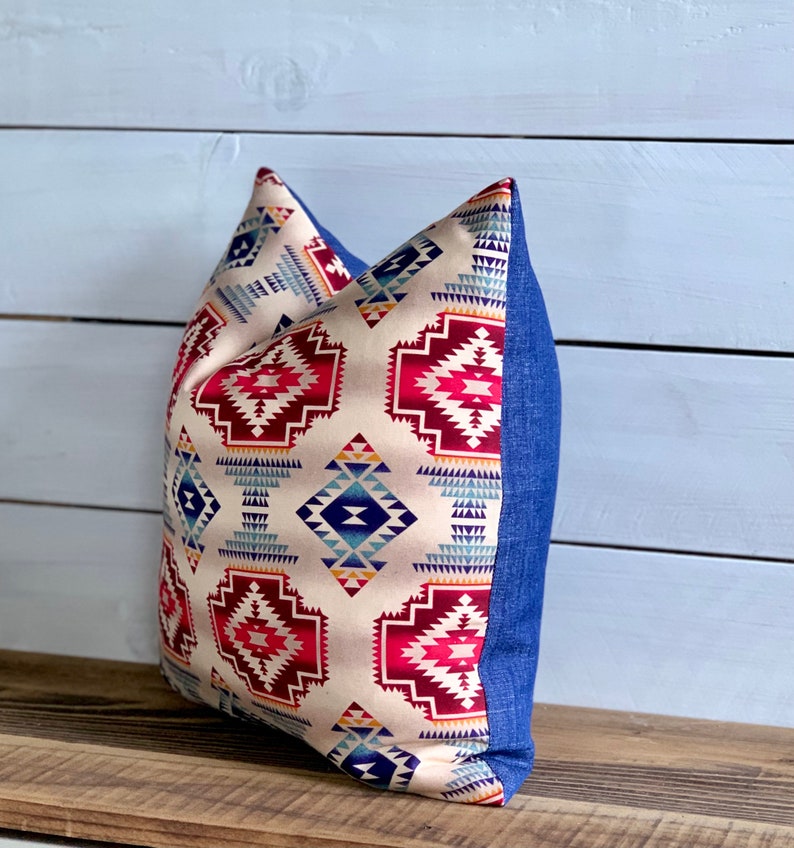 May include: A decorative pillow with a geometric pattern in shades of red, blue, and yellow on a cream background. The pillow has a denim blue border.