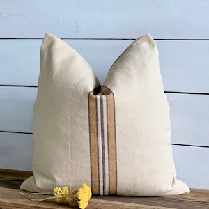 May include: A cream-colored pillow with brown and tan stripes down the center. The pillow is sitting on a wooden surface.