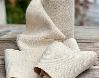 May include: A roll of natural-colored cotton webbing, a type of heavy-duty fabric tape, is shown on a wooden surface. The webbing is folded and rolled, revealing its texture and weave.