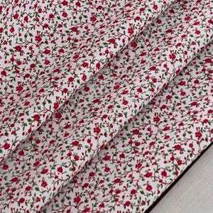 Puede incluir: Tela blanca con un patrón repetido de pequeñas rosas rojas y hojas verdes sobre finos tallos sinuosos. La tela está doblada, mostrando la continuidad del patrón y la textura del material. El diseño es un estampado floral.