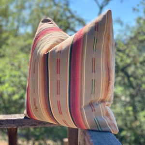 May include: A decorative pillow with a striped pattern in shades of tan, red, brown, and green. The pillow is resting on a wooden railing, with a blurred natural background.