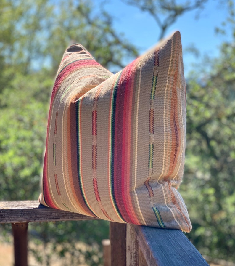 May include: A decorative pillow with a striped pattern in shades of tan, red, brown, and green. The pillow is resting on a wooden railing, with a blurred natural background.