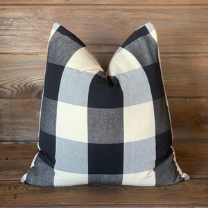 May include: A square pillow with a black, white, and gray buffalo check pattern. The pillow is made of a soft fabric and is filled with a fluffy material. The pillow is a decorative item for a bed or couch.