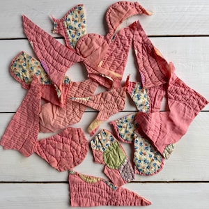 May include: A collection of pink quilted fabric pieces in various shapes and sizes, some with floral patterned accents. The pieces are arranged on a white wooden surface, showcasing a handmade craft project.