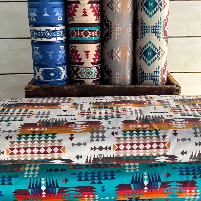 May include: Four rolls of fabric with geometric patterns in shades of blue, red, white, and brown. The fabric is stacked on a wooden crate. The fabric is folded and has a geometric pattern.