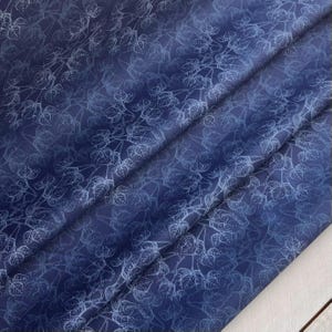 May include: Dark blue fabric with a repeating pattern of white leaf outlines. The fabric is folded, creating diagonal lines across the surface. The leaves are detailed, with visible veins and varying sizes.