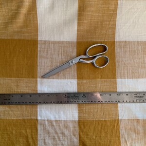 May include: A silver pair of scissors and a metal ruler on a white and mustard yellow checkered fabric.