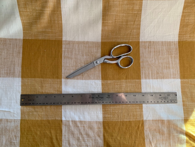 May include: A silver pair of scissors and a metal ruler on a white and mustard yellow checkered fabric.
