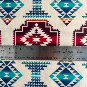 May include: A white fabric with a repeating pattern of colorful geometric shapes. The pattern features shades of red, blue, turquoise, and gold. A ruler is placed on the fabric, showing the scale in inches.