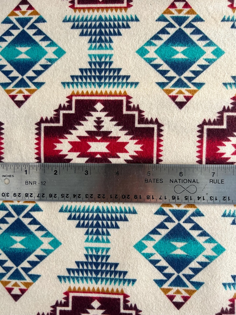 May include: A white fabric with a repeating pattern of colorful geometric shapes. The pattern features shades of red, blue, turquoise, and gold. A ruler is placed on the fabric, showing the scale in inches.