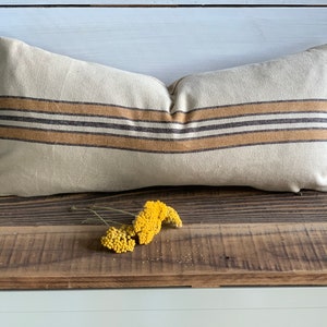 May include: A long, rectangular pillow with a beige linen cover and brown and tan striped pattern. The pillow is sitting on a wooden surface with a small bouquet of yellow flowers in front of it.