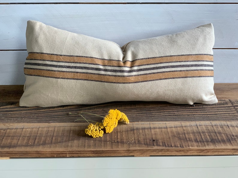 May include: A long, rectangular pillow with a beige linen cover and brown and tan striped pattern. The pillow is sitting on a wooden surface with a small bouquet of yellow flowers in front of it.
