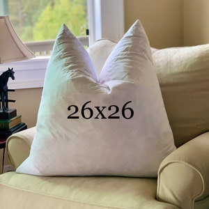 May include: A white pillow with a 26x26 inch size label on it, sitting on a light beige armchair.