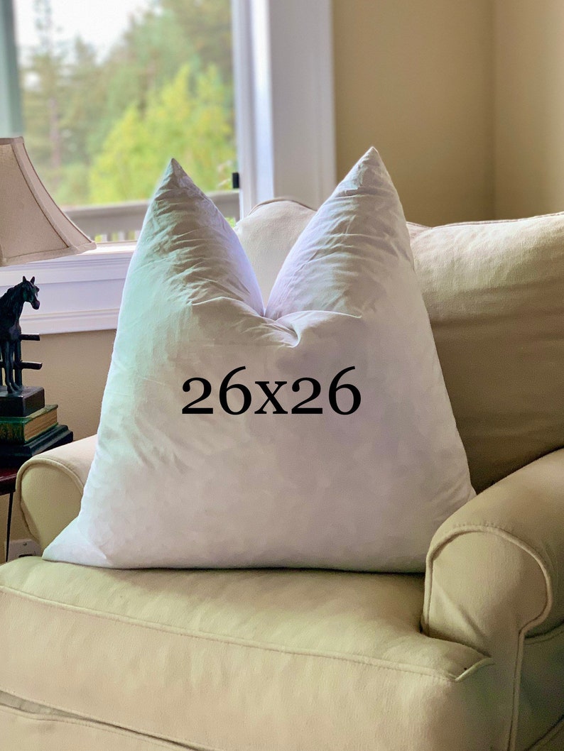 May include: A white pillow with a 26x26 inch size label on it, sitting on a light beige armchair.