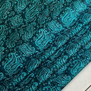 May include: Dark teal fabric with a paisley pattern in a lighter teal color. The fabric is folded, showing the intricate design and texture. The paisley design is a classic, elegant pattern.