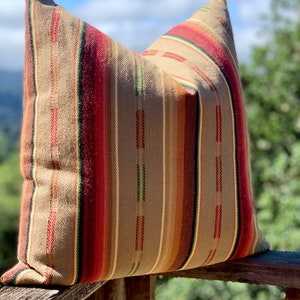 May include: A decorative pillow with a woven fabric cover. The pillow features vertical stripes in shades of brown, red, orange, and green. The pillow is resting on a wooden railing, with a blurred background of trees and sky.