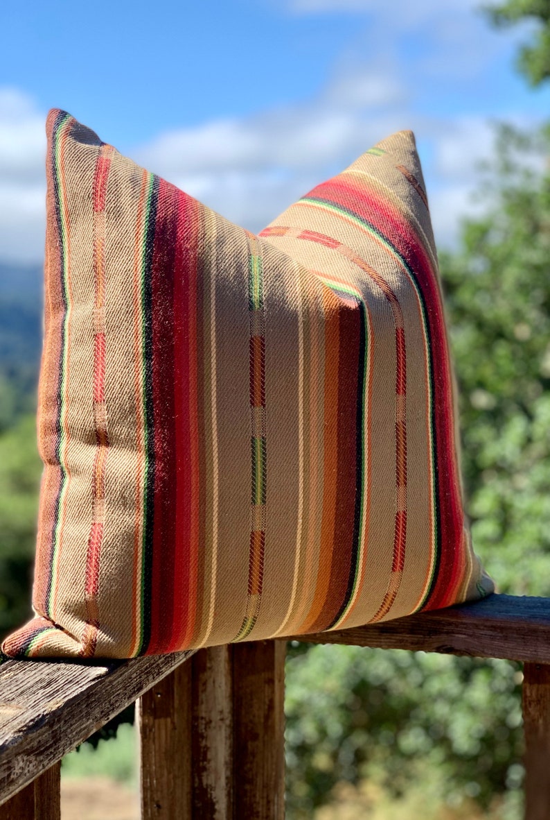 May include: A decorative pillow with a woven fabric cover. The pillow features vertical stripes in shades of brown, red, orange, and green. The pillow is resting on a wooden railing, with a blurred background of trees and sky.